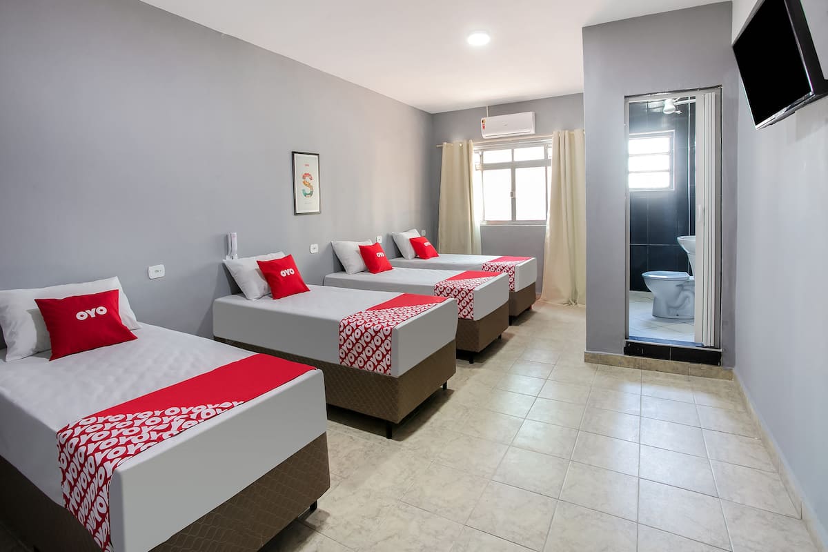 Una camera d'albergo con tre letti, ognuno con copripiumino bianco e rosso e un cuscino rosso con il logo "oyo". Si nota una lampada a soffitto, un condizionatore d'aria e un televisore montato a parete.