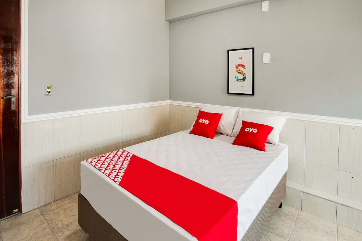 Un letto rifatto con cura, con biancheria rossa e bianca, un quadro appeso al muro e una porta alla sinistra.