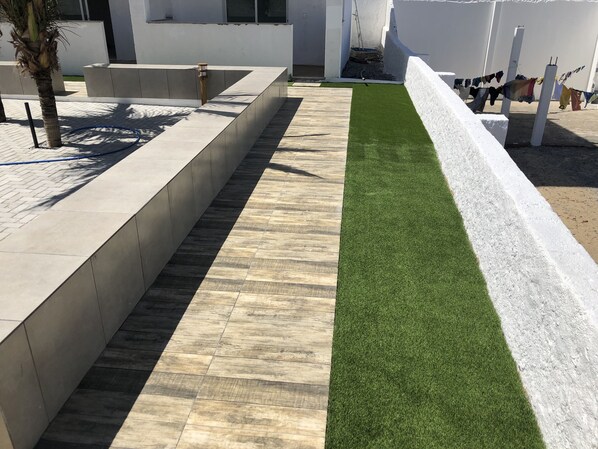 A pathway with artificial grass and tiled flooring, bordered by white walls and a palm tree. - Vilas Blancas Apartamentos (Caucaia)