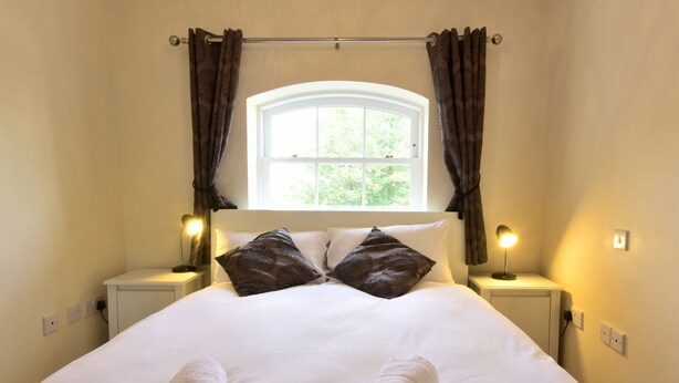 A neatly made bed with white linens and two decorative pillows, a bedside lamp, and a window with curtains.