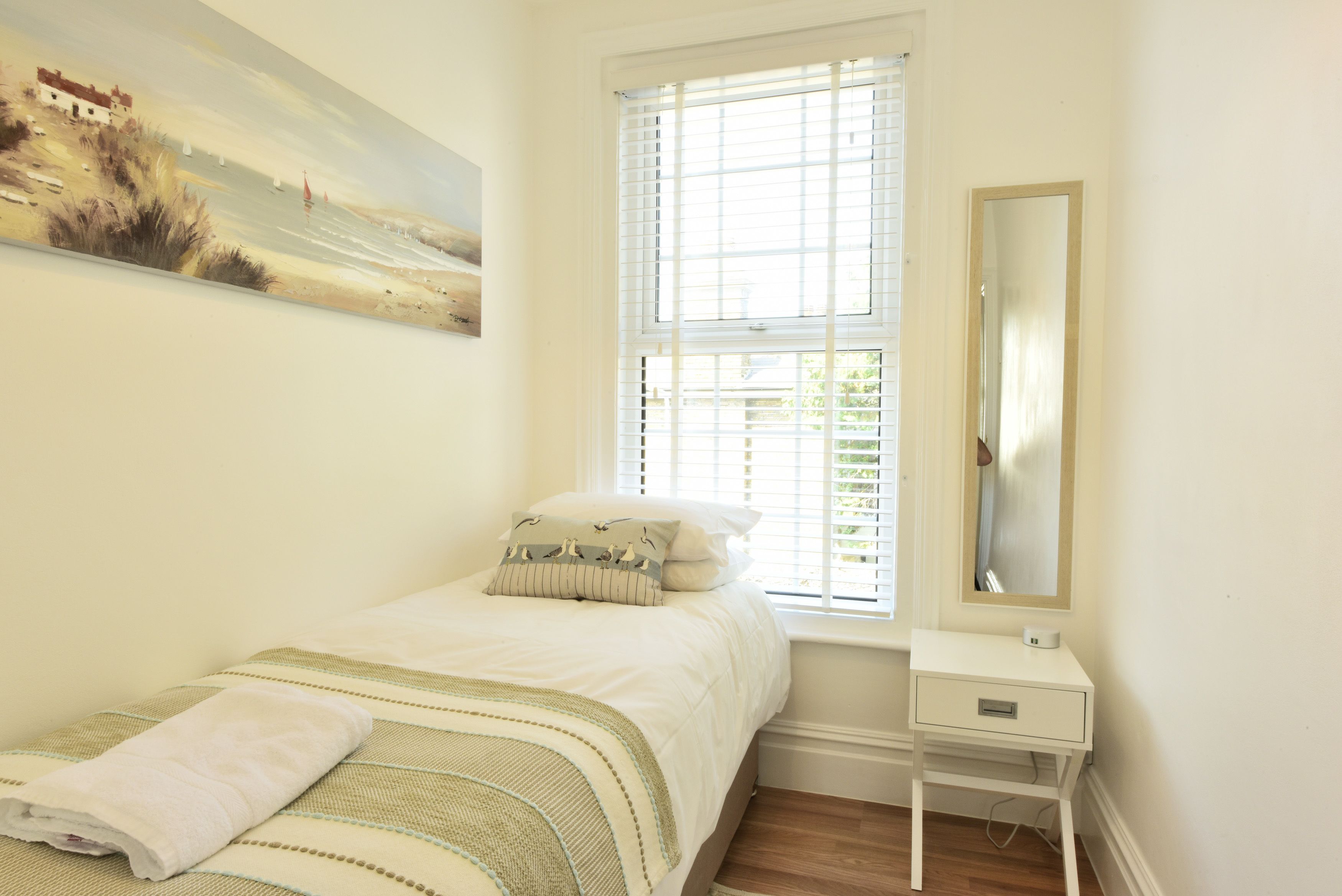 A bedroom with a bed, a bedside table, a window with blinds, and a painting on the wall.