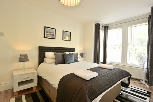 A neatly made bed with a dark headboard, white bedding, and a folded towel on the footboard. There are two bedside tables with lamps, a window with curtains, and a radiator. - Grosvenor House (Margate)