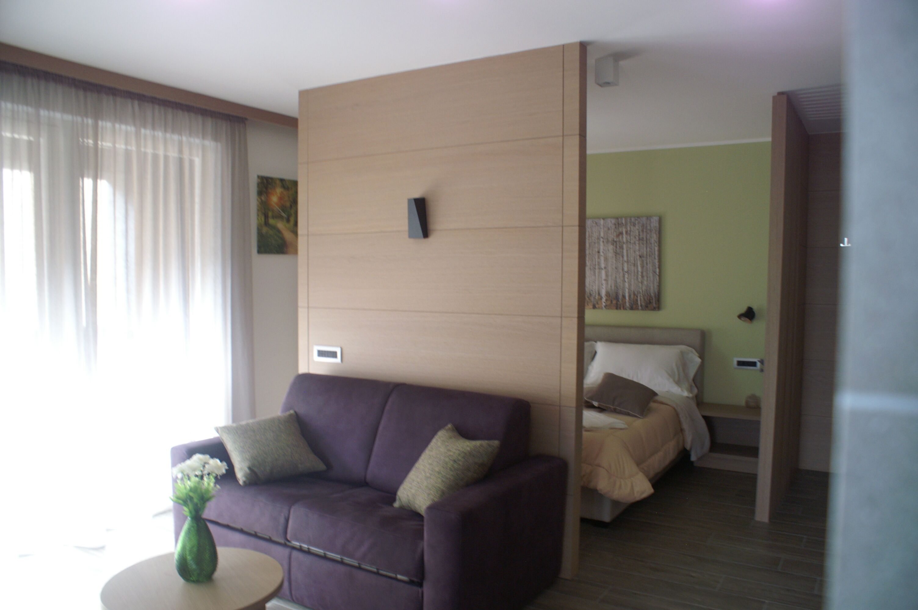 A modern hotel room with a purple sofa, a small round table with a vase, a bed with a wooden headboard, and a green wall with artwork.