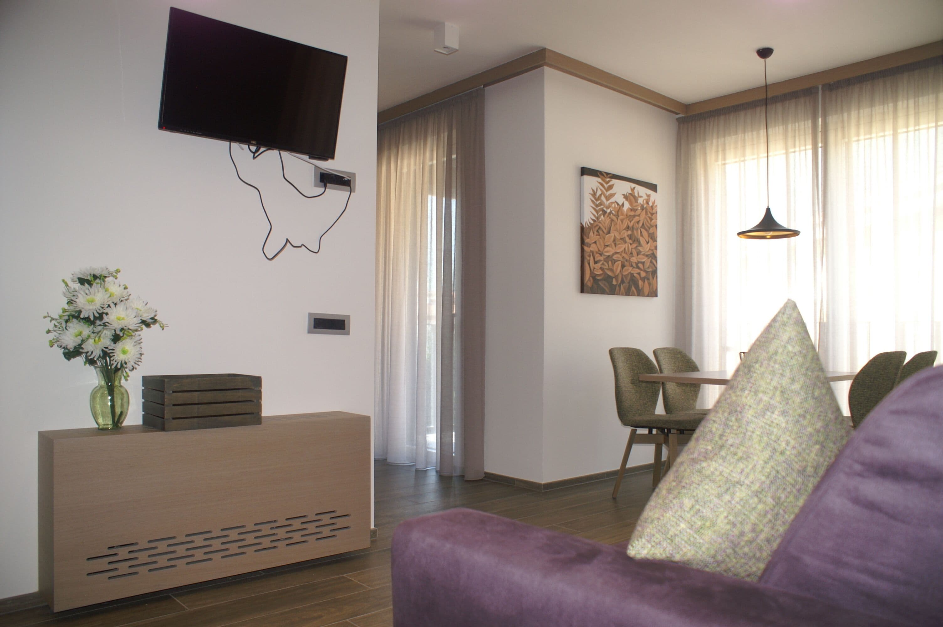 Un salon moderne avec un téléviseur à écran plat fixé au mur, une console en bois sur laquelle est posé un vase de fleurs, un canapé violet et un coin repas avec une table et des chaises.