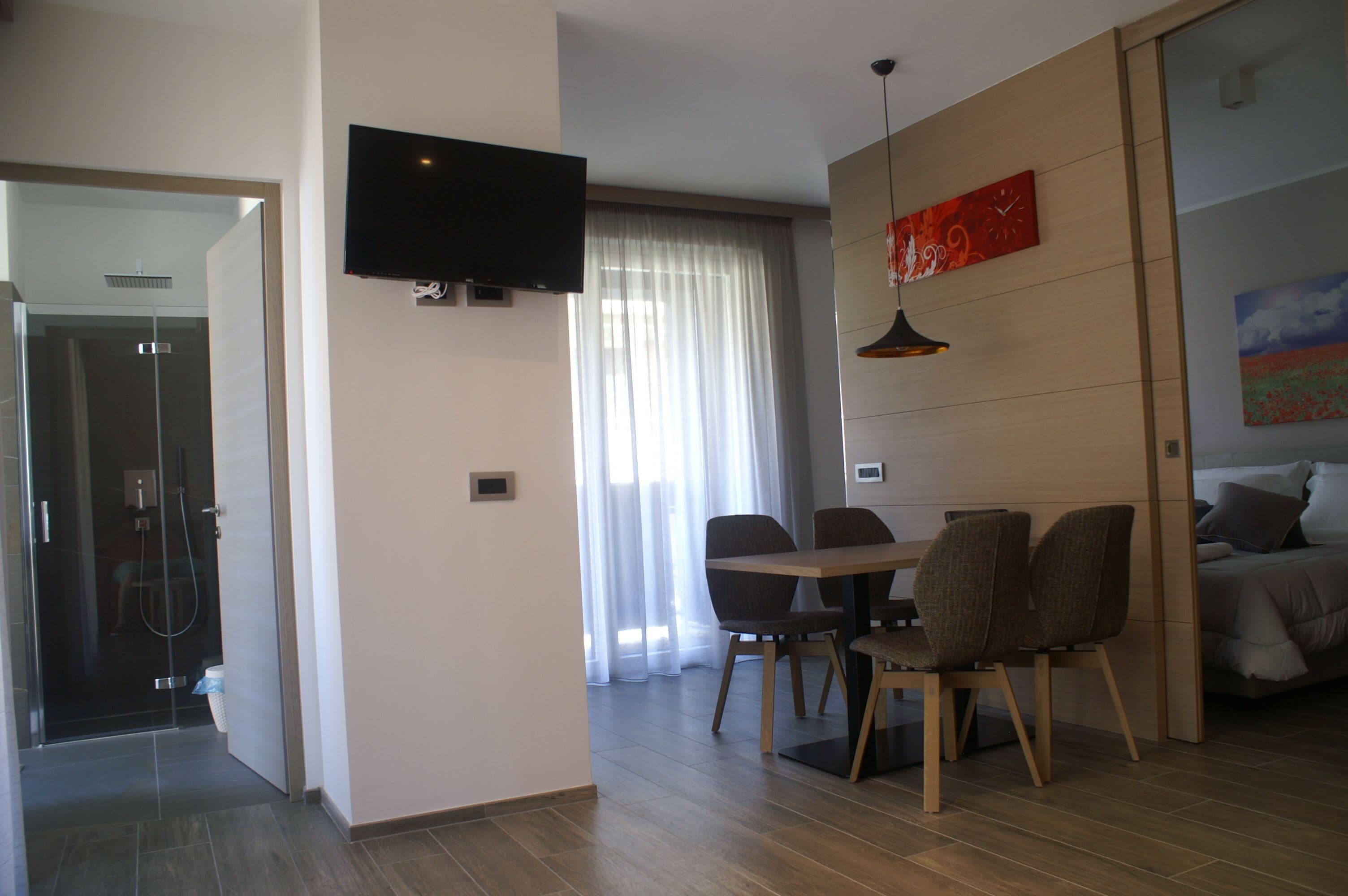 Una habitación de hotel moderna con ducha, un área de comedor con mesa y sillas, un televisor montado en la pared y una cama con un cuadro encima.