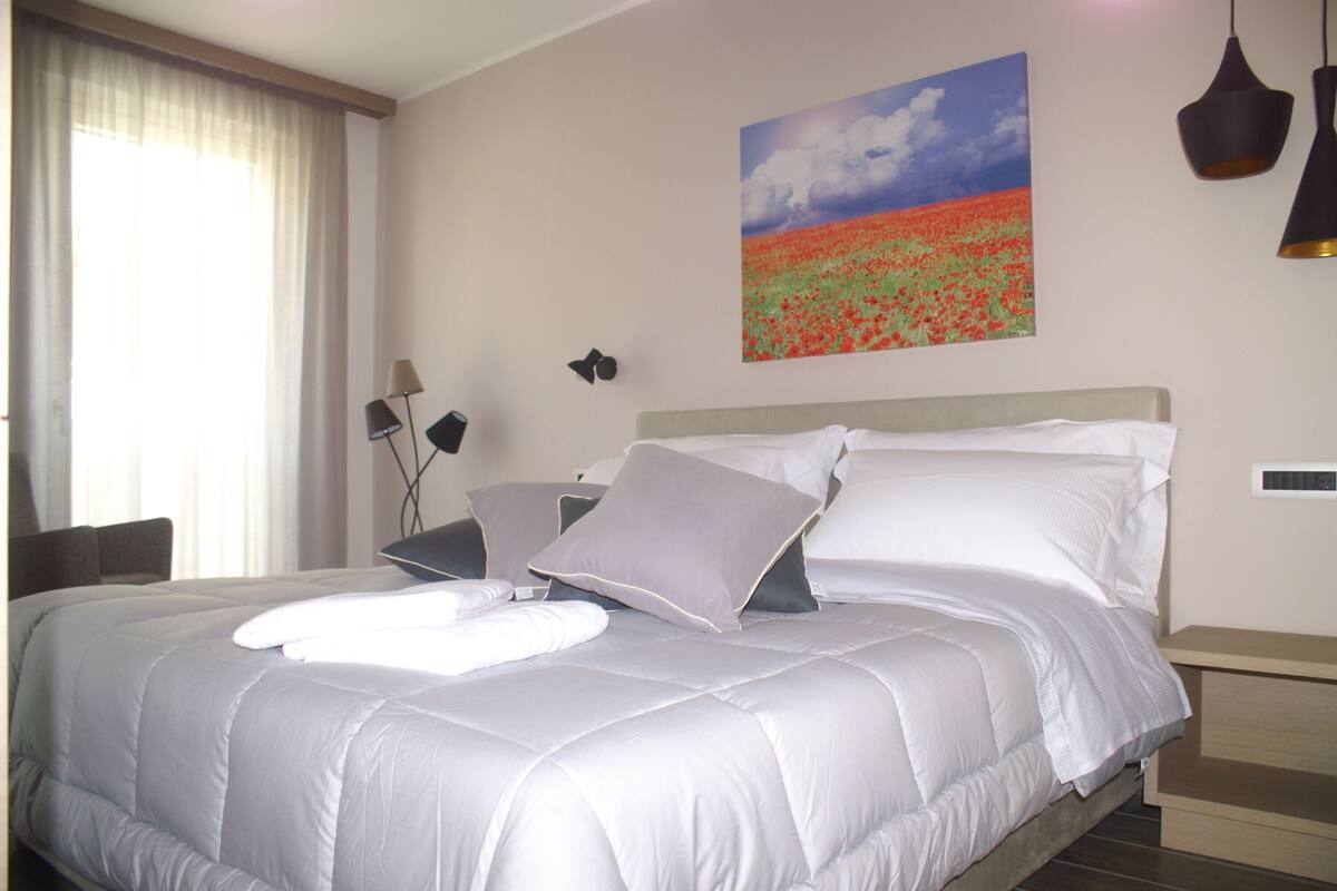 A neatly made bed with white linens and pillows, a painting of a field on the wall, and a lamp on a bedside table.
