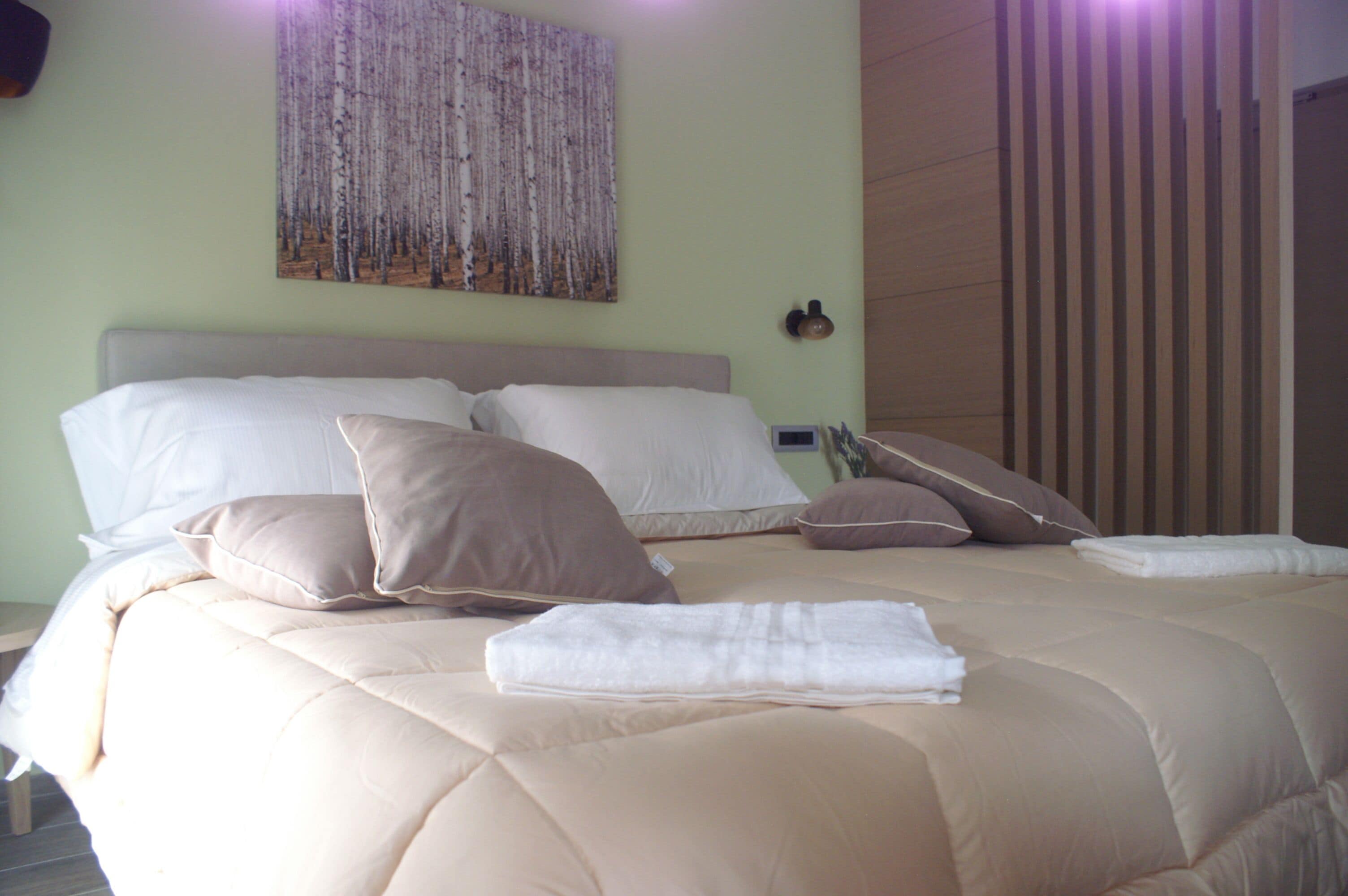 Una cama bien hecha con un edredón color beige y varias almohadas, una obra de arte en la pared que representa un bosque, y un panel de madera al lado derecho.