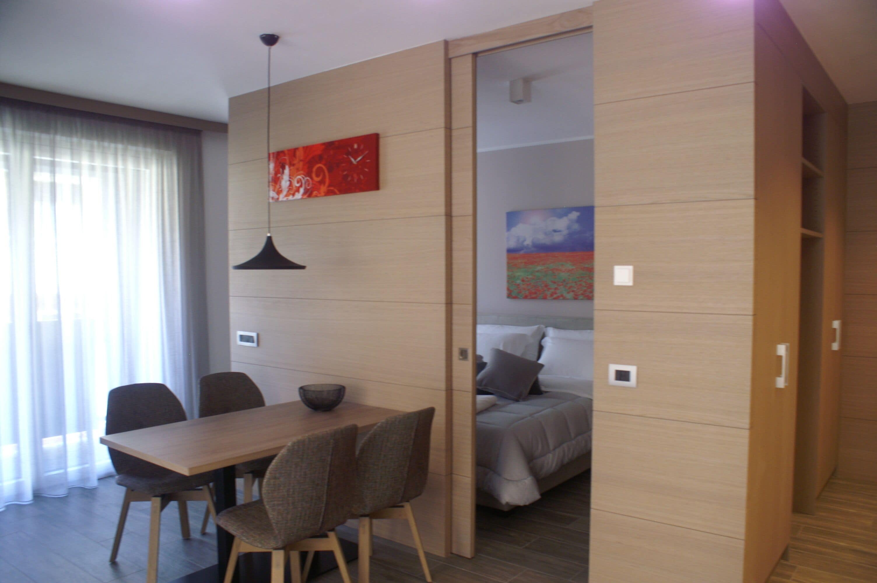 Una habitación moderna con una mesa de madera, sillas, una cama con un cuadro y una puerta corredera.