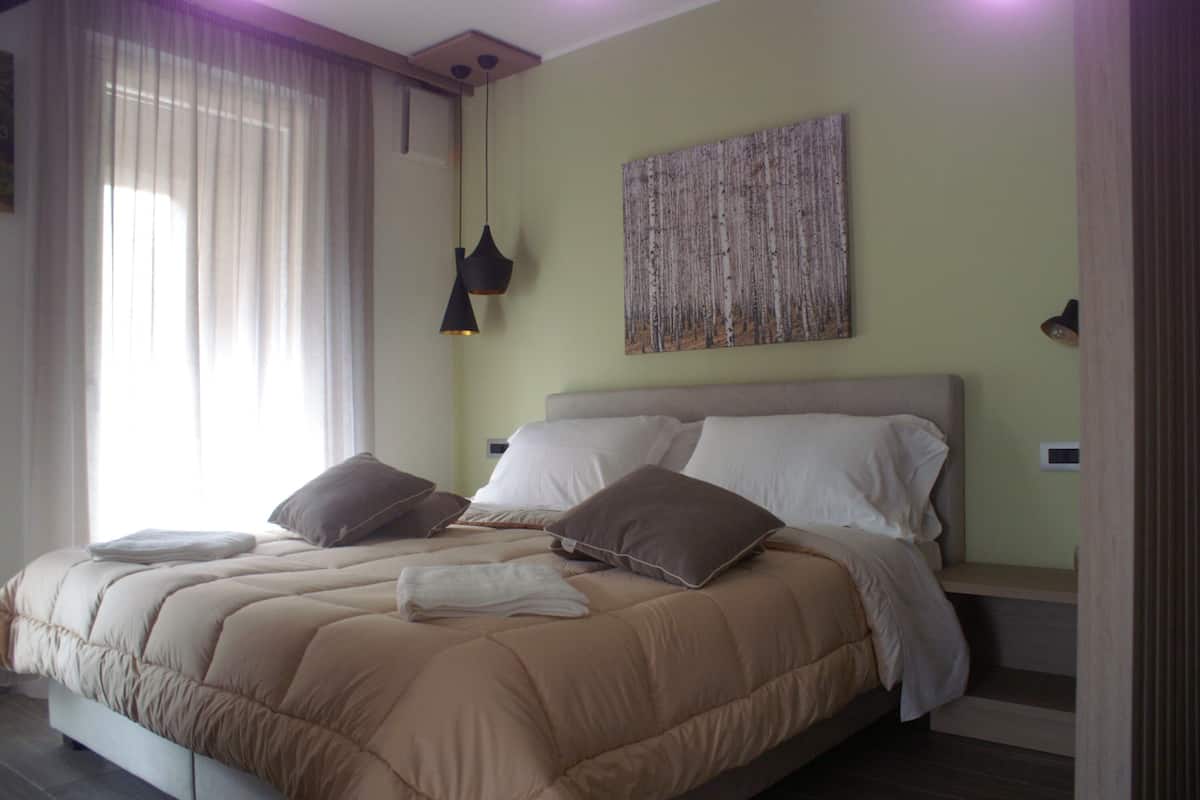A neatly made bed with a beige duvet and pillows, a large abstract painting on the wall, and a hanging pendant light.