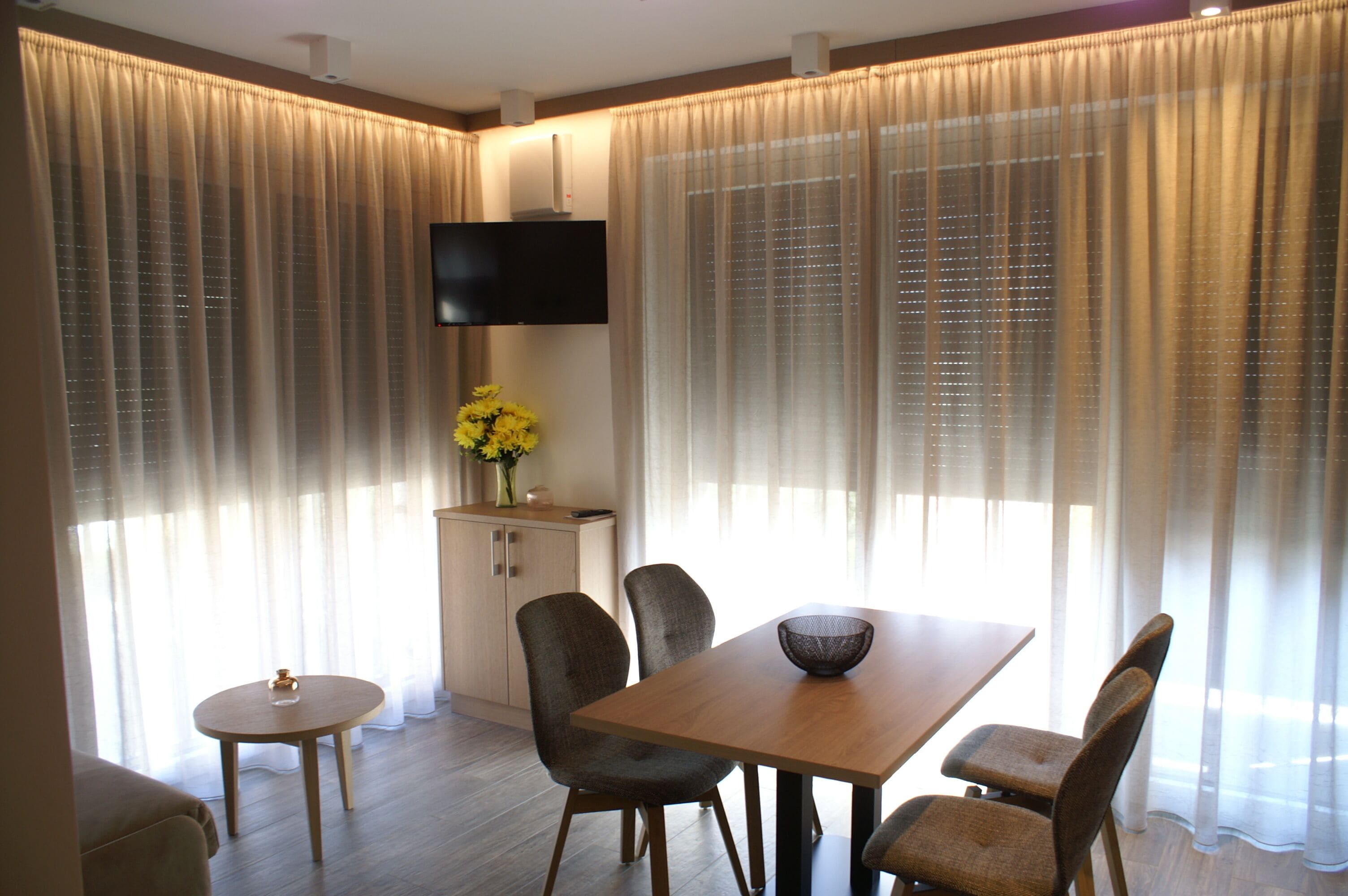 Un espace repas moderne avec une table et des chaises en bois, une petite table ronde, un vase avec des fleurs jaunes et un téléviseur à écran plat fixé au mur.