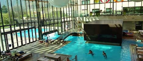 An indoor swimming pool with clear glass walls, lounge areas, and a view of the mountains.