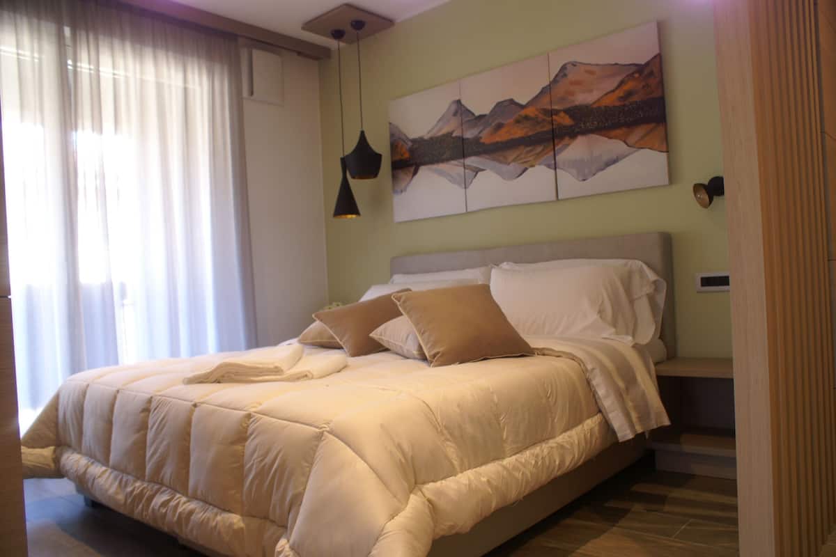 A neatly made bed with a beige comforter and pillows, a painting of mountains on the wall, and a window with sheer curtains.