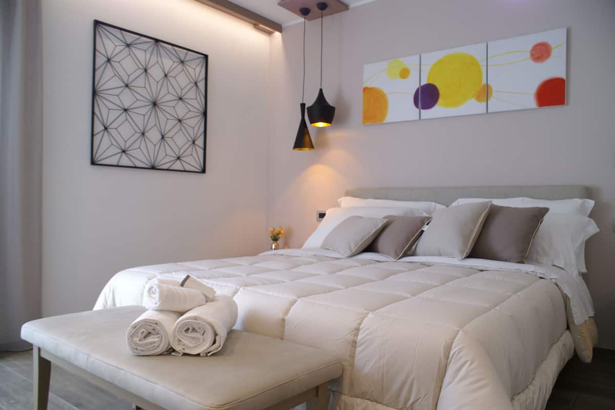 A neatly made bed with a white comforter and pillows, a bedside table with folded towels, and a wall art piece above the bed.