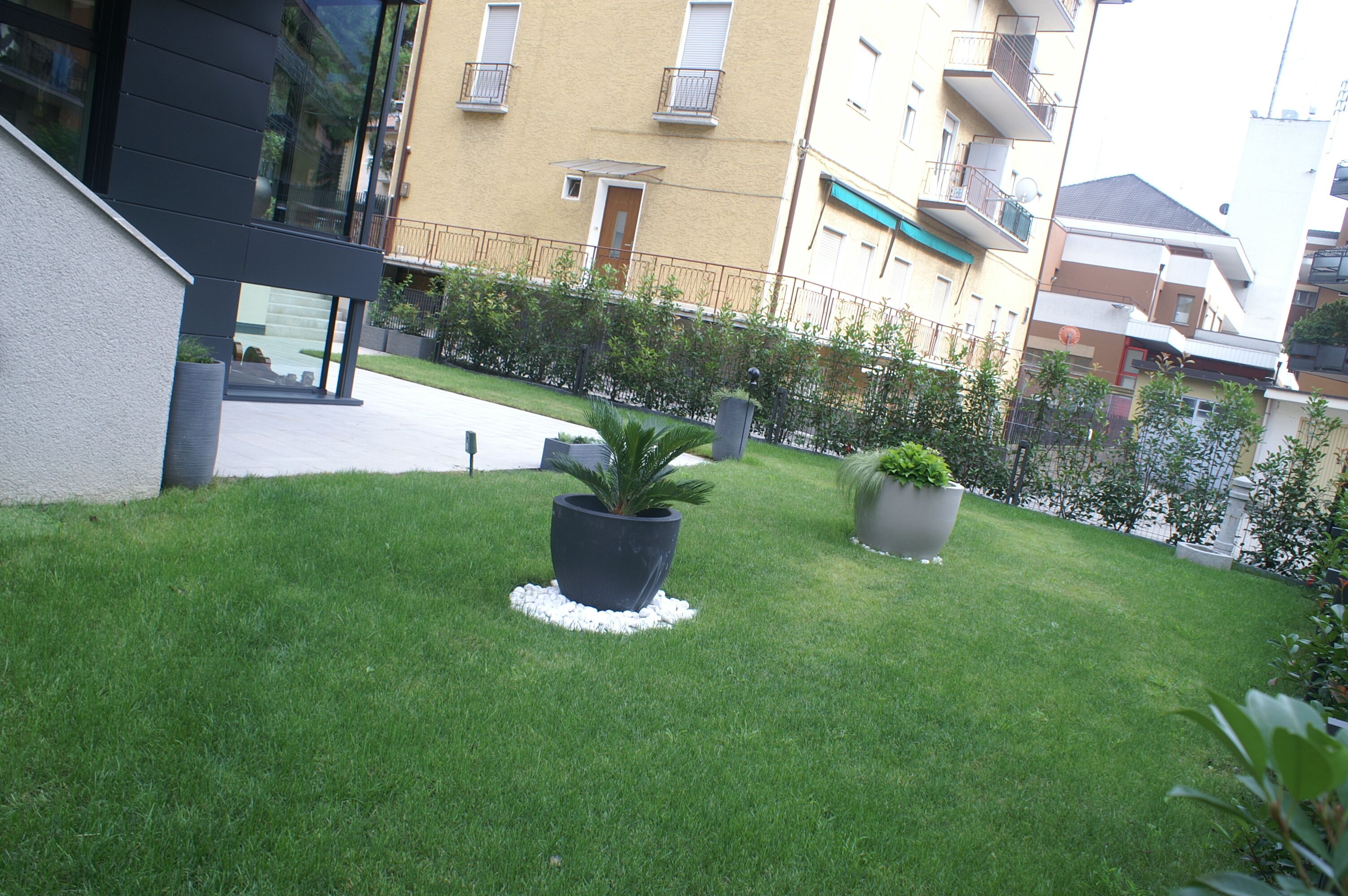 A well-maintained grassy area with two large potted plants, a modern building with balconies, and a clear view of the surrounding residential buildings.