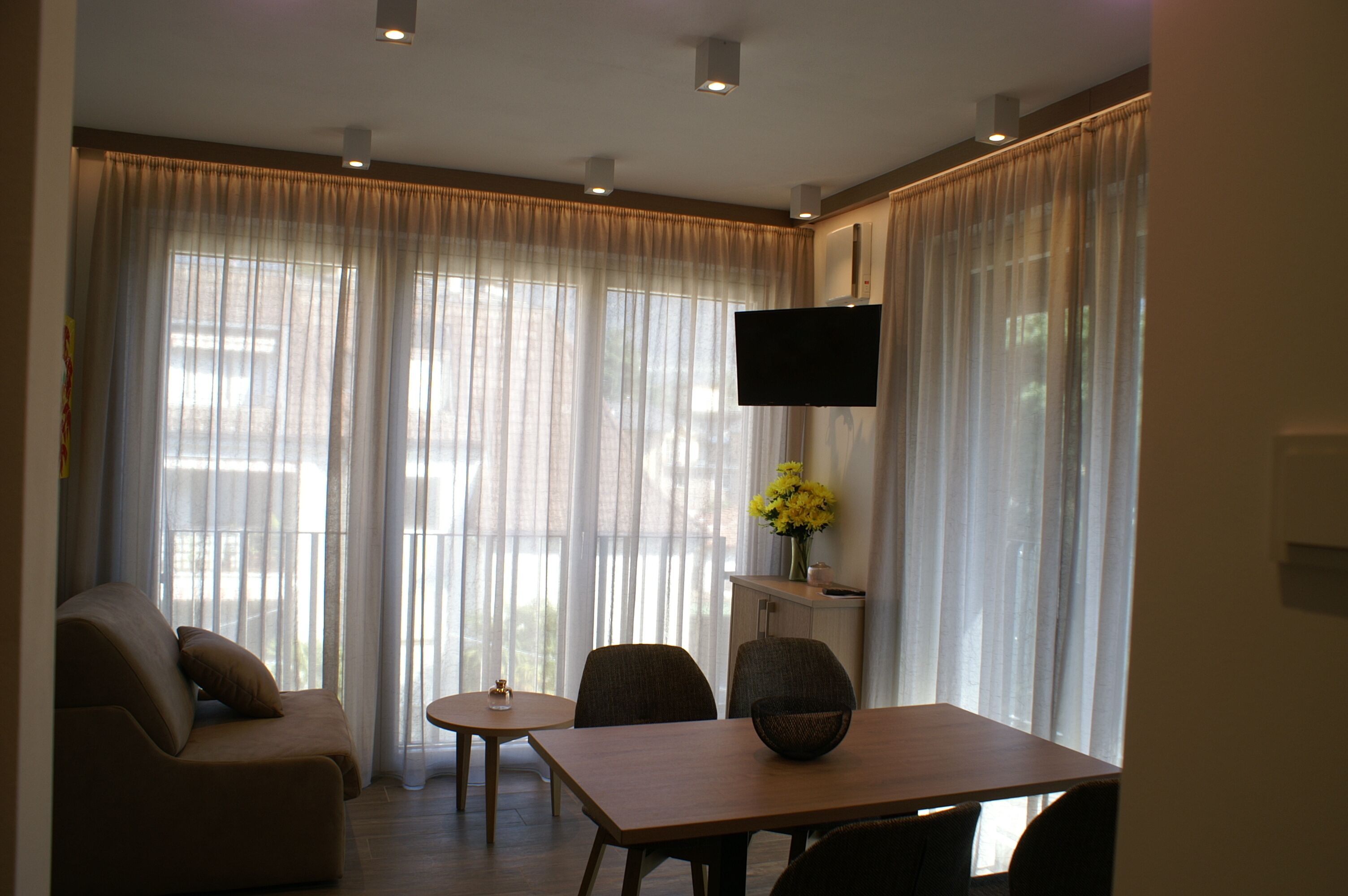 A modern living room with a sofa, a dining table, and a flat-screen TV mounted on the wall.