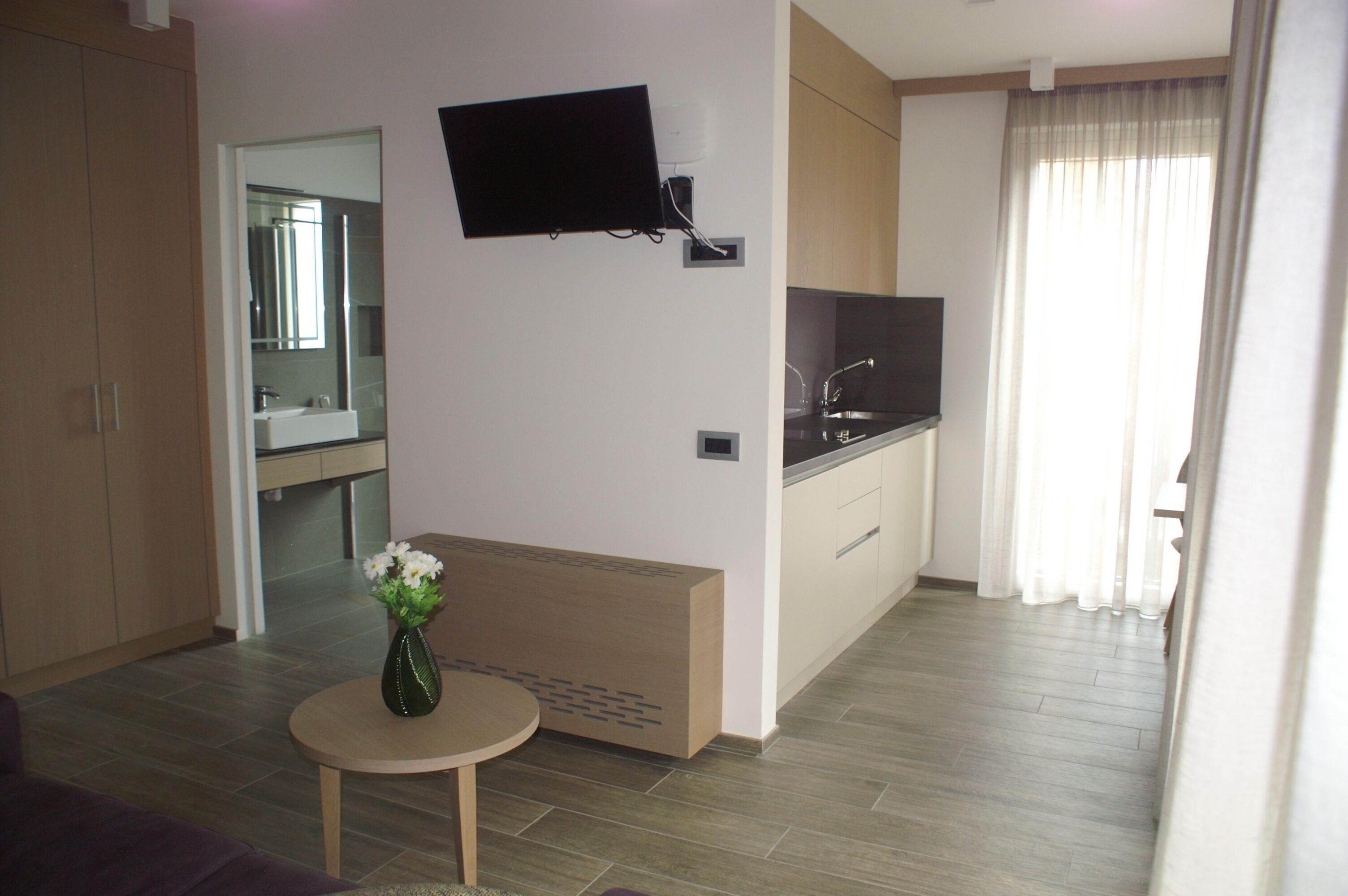 A modern hotel room with a kitchenette, a flat-screen TV mounted on the wall, a small round table with a vase of flowers, and a large window with sheer curtains.