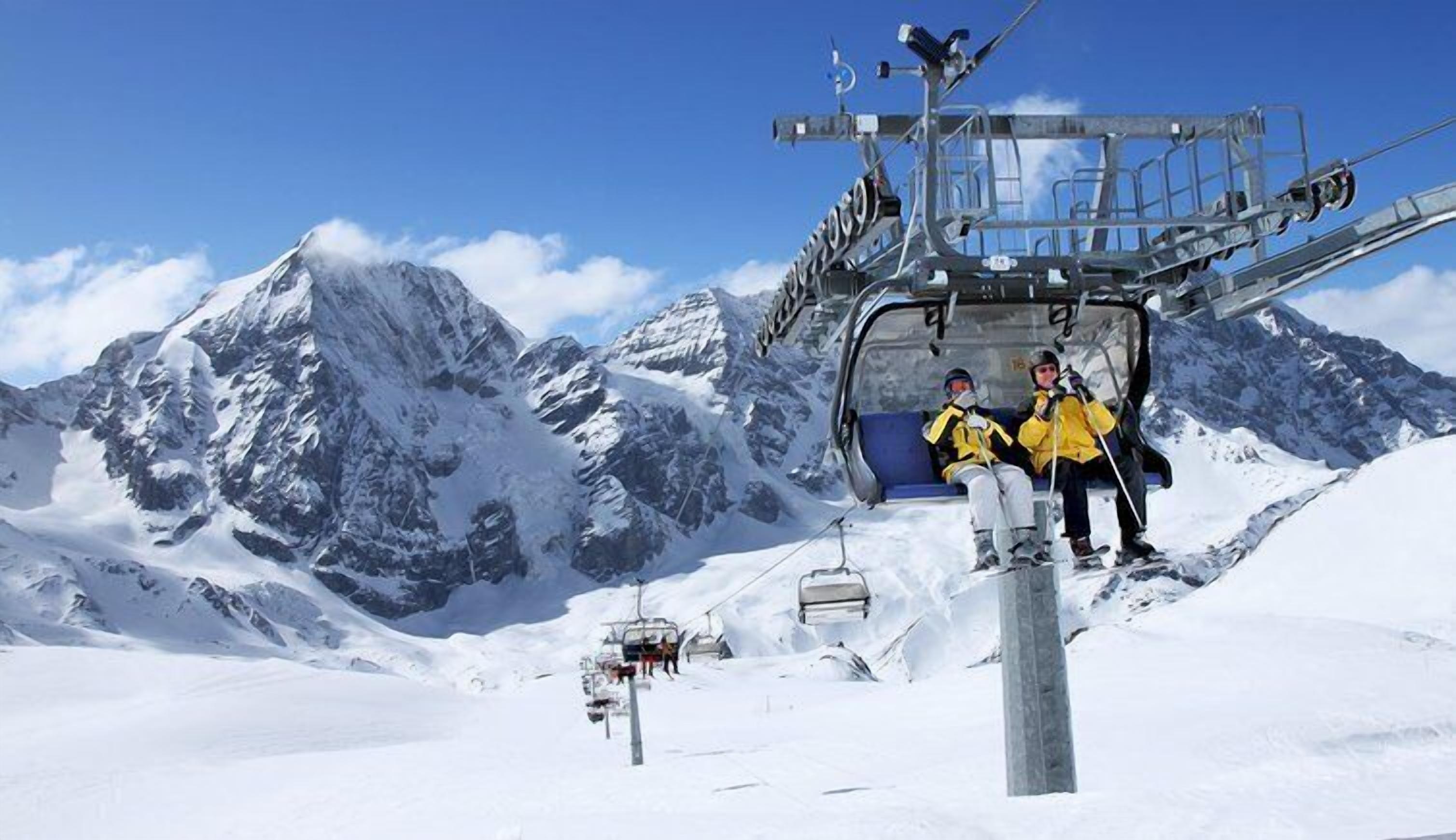 Un télésiège avec des passagers, des montagnes enneigées en arrière-plan et un ciel dégagé.