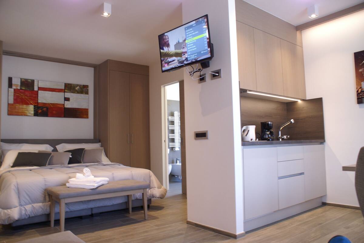 A modern hotel room with a bed, a small desk, a television mounted on the wall, and a kitchen area with a sink and countertop.