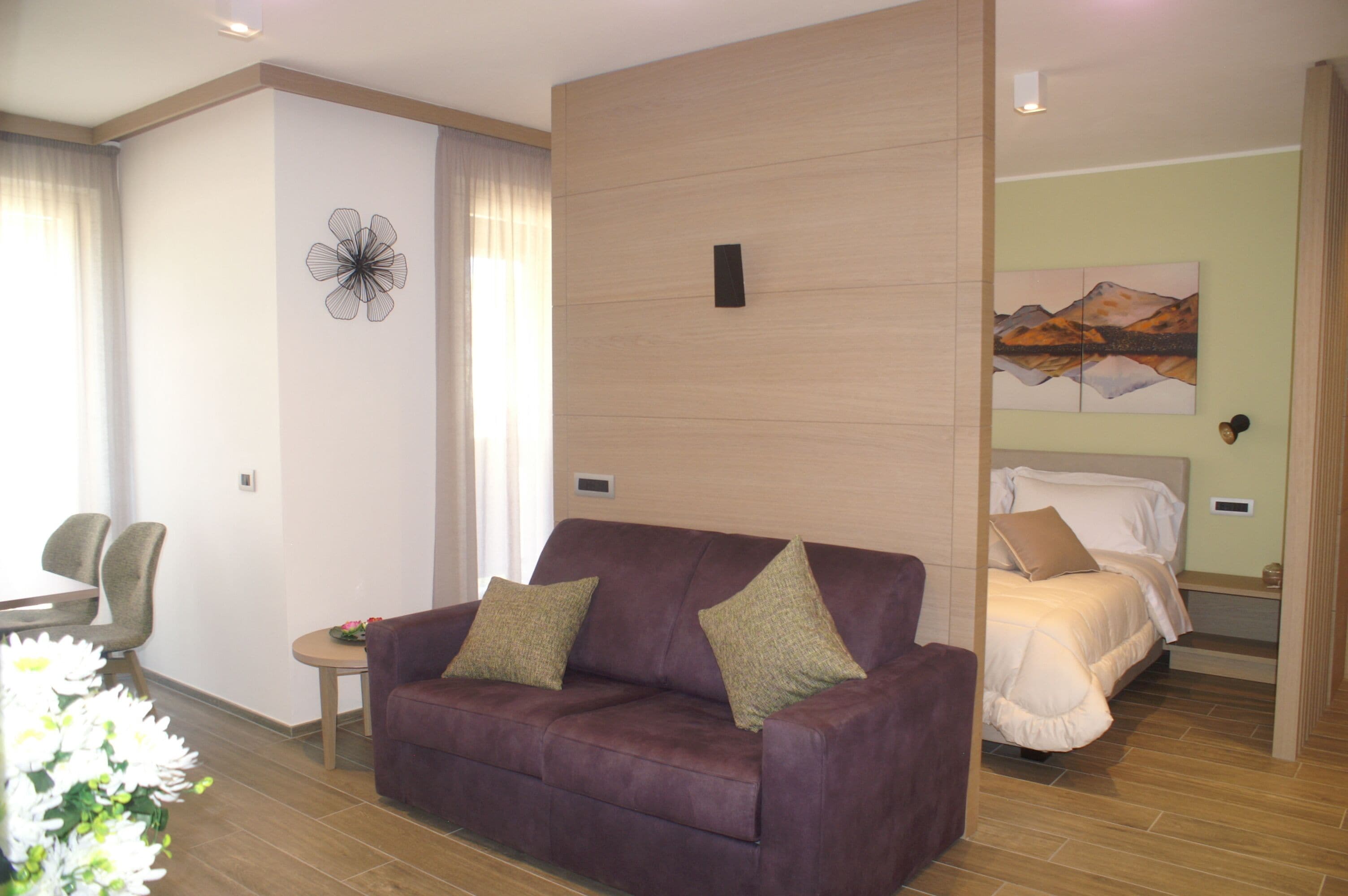 Una habitación de hotel moderna con un sofá morado, una cama con ropa de cama blanca, una obra de arte de montañas y una partición de madera.