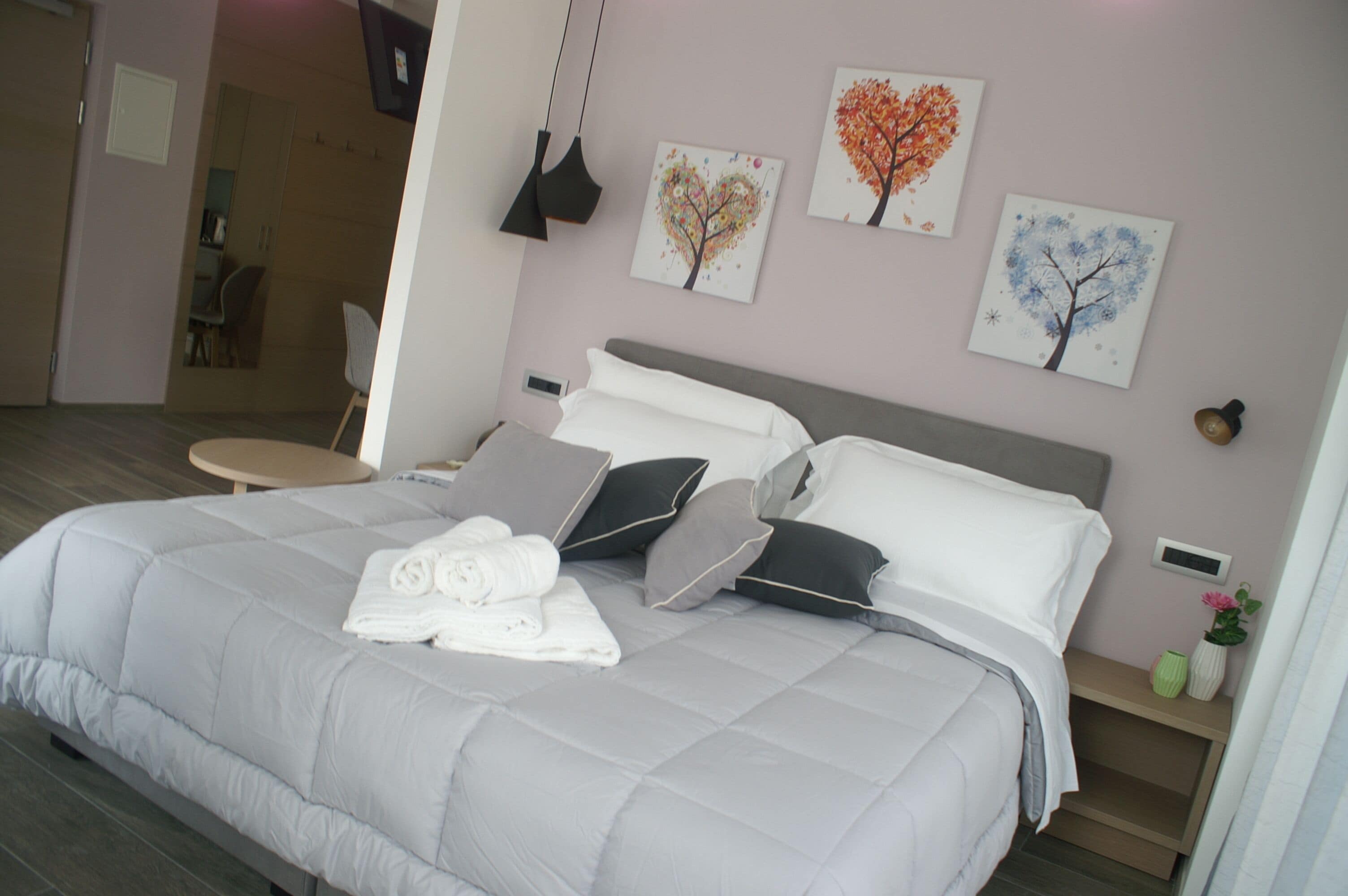 A neatly made bed with pillows and a rolled towel, a bedside table with a vase, and wall art depicting trees.