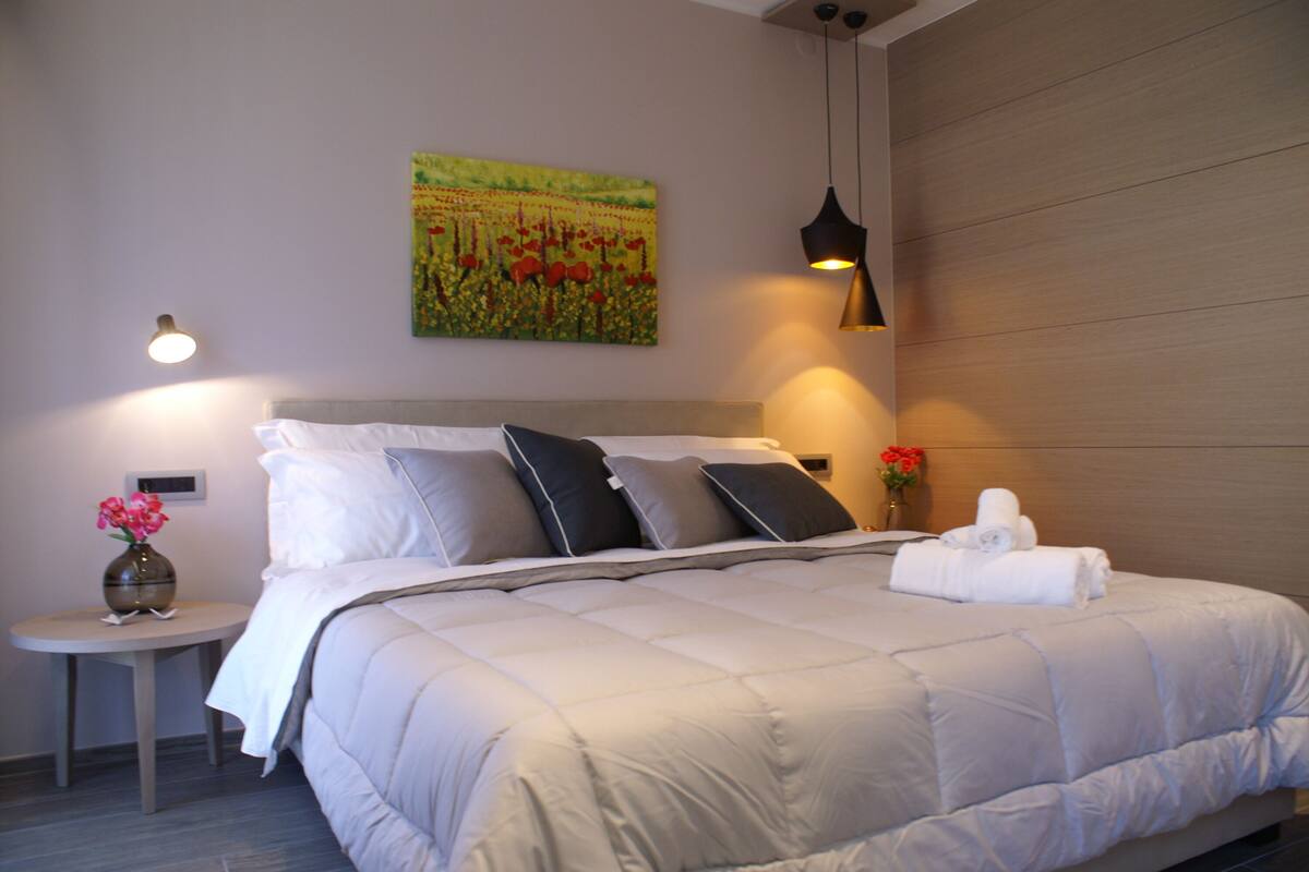 A neatly made bed with a headboard, a bedside table with a vase of flowers, and a wall-mounted painting.