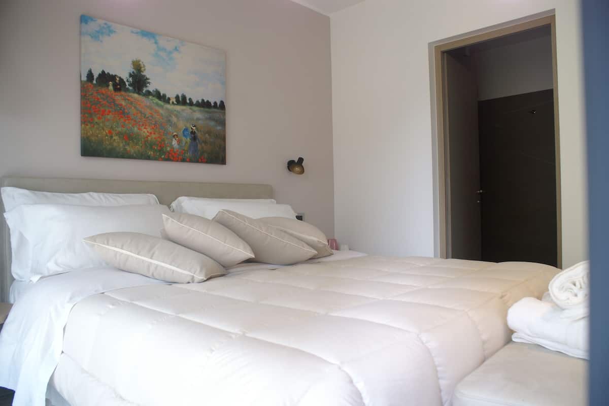 A neatly made bed with white linens and pillows, a painting of a landscape on the wall, and a door to the right.