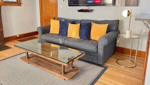 Ein modernes Wohnzimmer mit einem grauen Sofa, gelben und blauen Kissen, einem Glastisch und einem flachbildfernseher an der Wand.