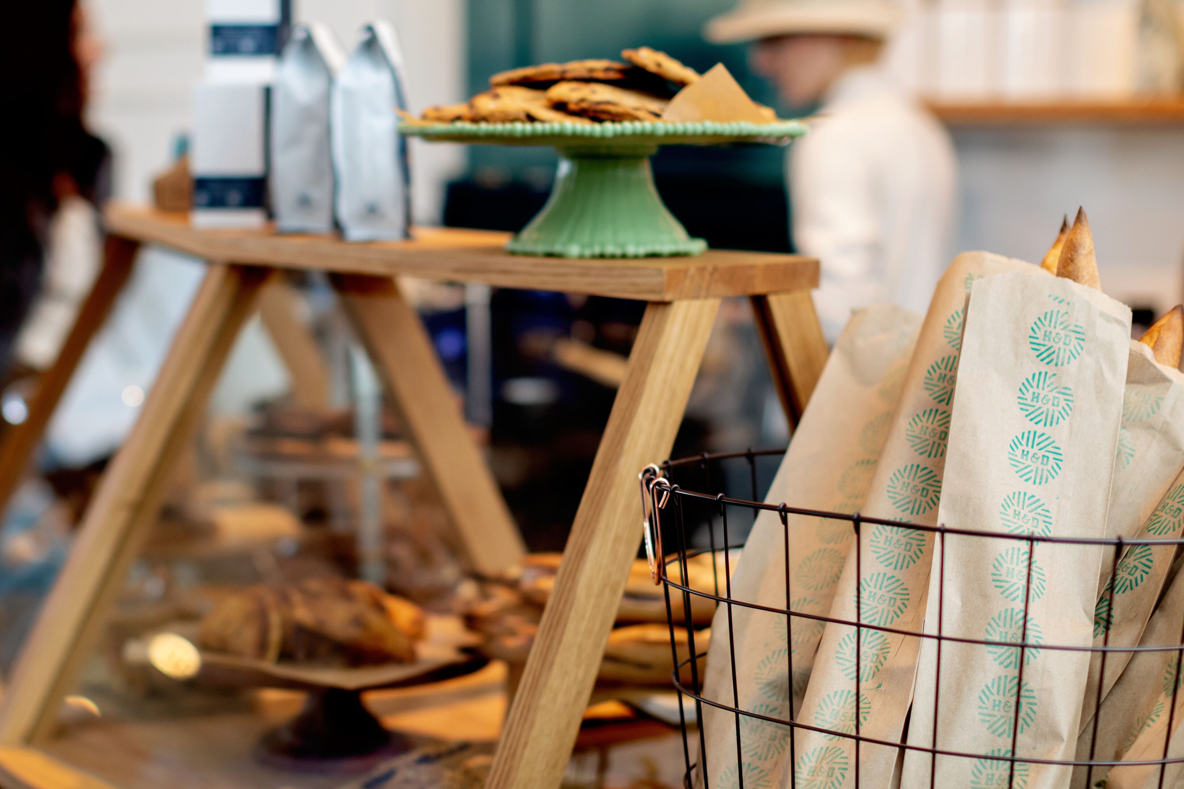 A display of baked goods on a wooden stand with a green base, a wire basket with paper bags, and a blurred background suggesting a bakery or cafe setting.