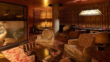 Une pièce comprenant une table de billard, deux fauteuils, une table basse en verre, une lampe et un tableau encadré représentant des oiseaux.