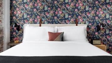 A neatly made bed with white linens and a single decorative pillow, set against a floral wallpaper backdrop.