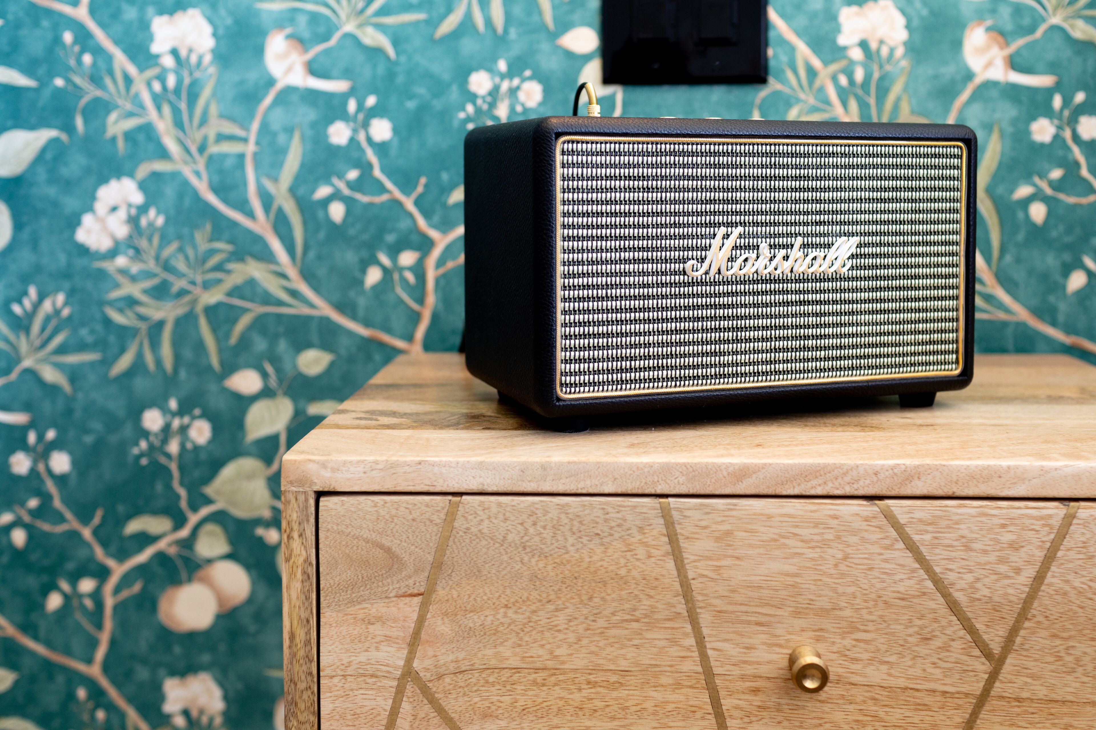 A Marshall amplifier on a wooden cabinet with a floral wallpaper background.