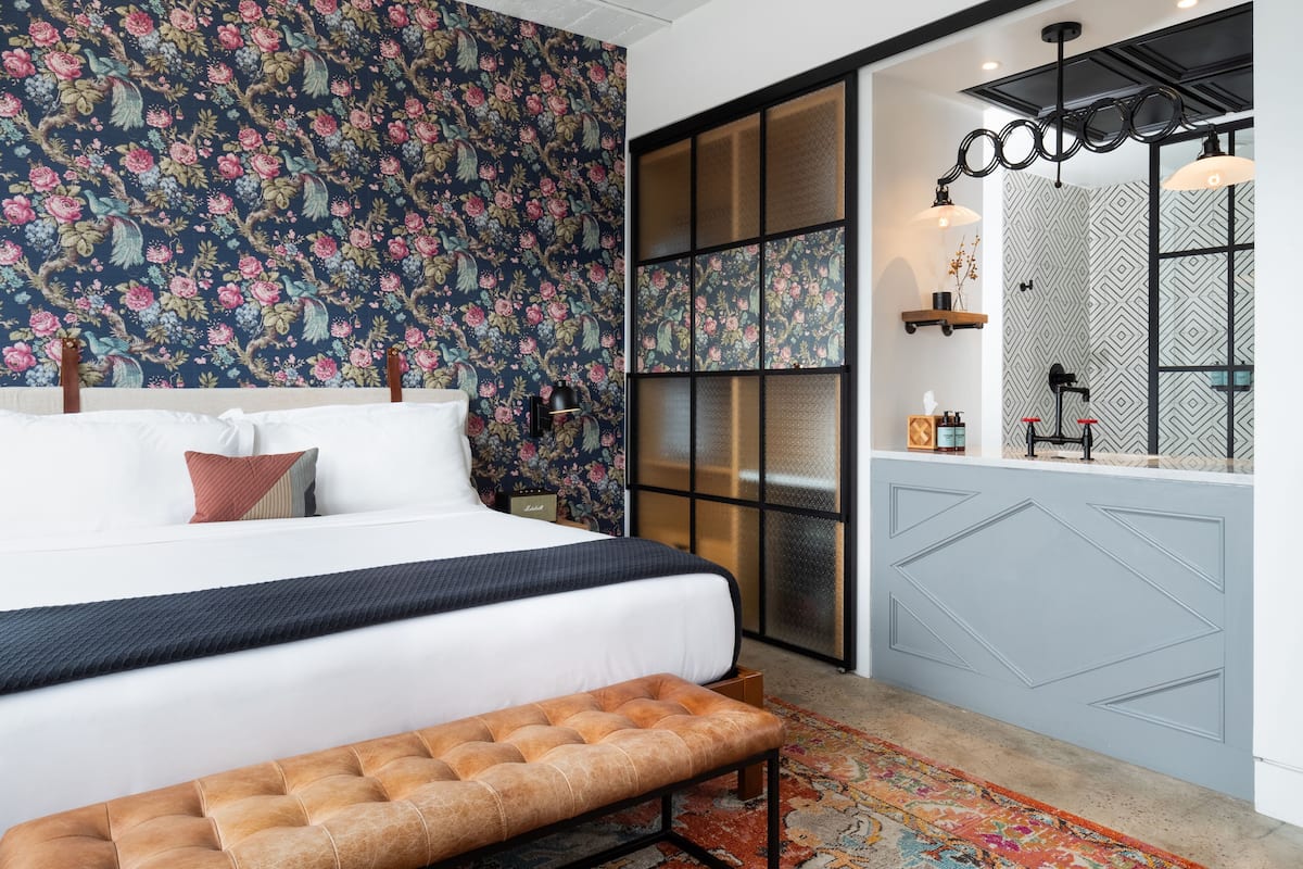 Ein Schlafzimmer mit einem großen Bett, einer Bank, einer Wand mit Muster, einer Schiebetür und einem Badezimmer mit Spiegel und Beleuchtung.