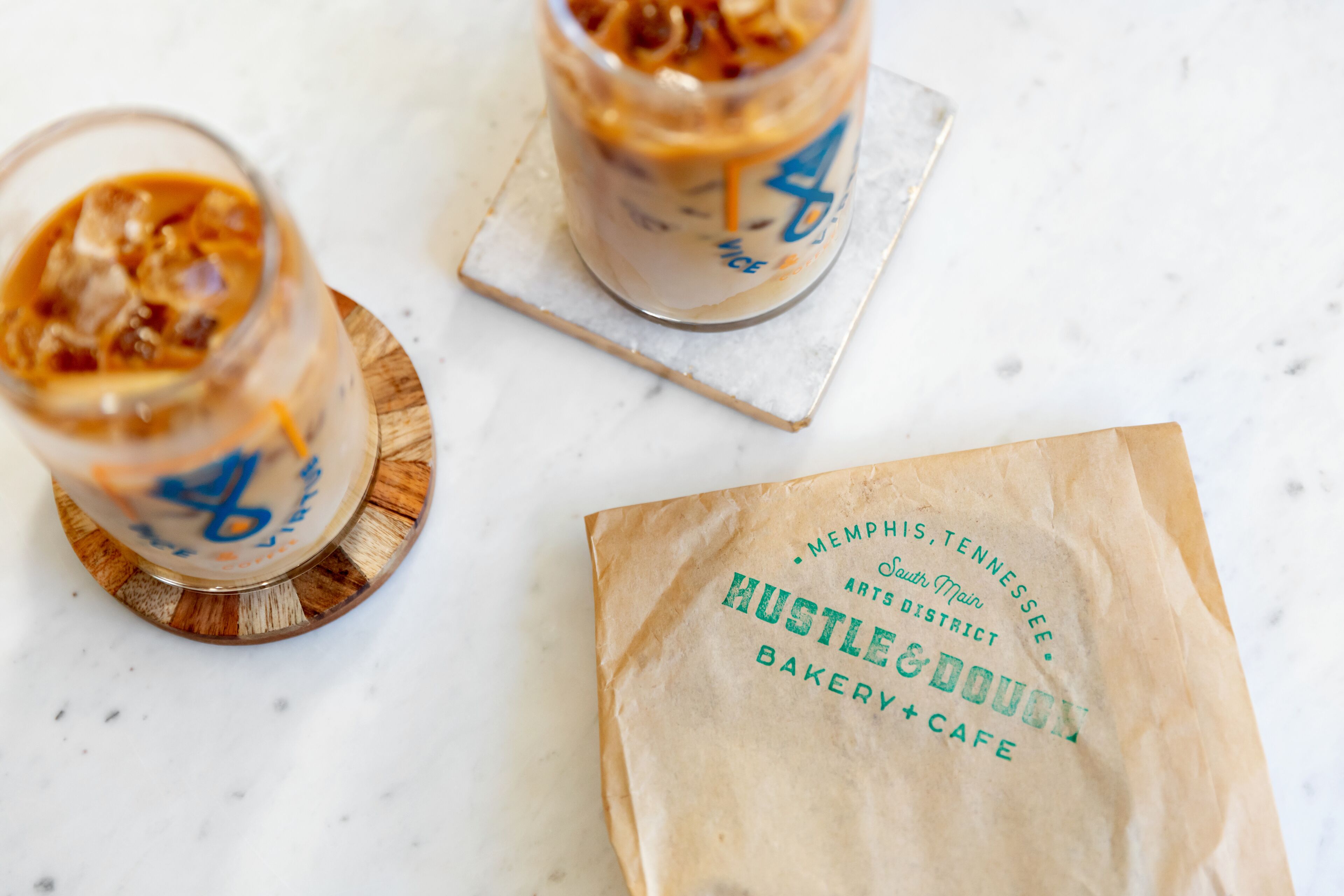Dos bebidas heladas sobre posavasos con una bolsa de papel que dice 'Hustle & Doughy Bakery & Cafe'.