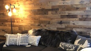 Ein Zimmer mit einer Holzwand, einem schwarzen Sofa mit gemusterten Kissen, einem Holztisch und einer Stehlampe mit drei Leuchtmitteln.