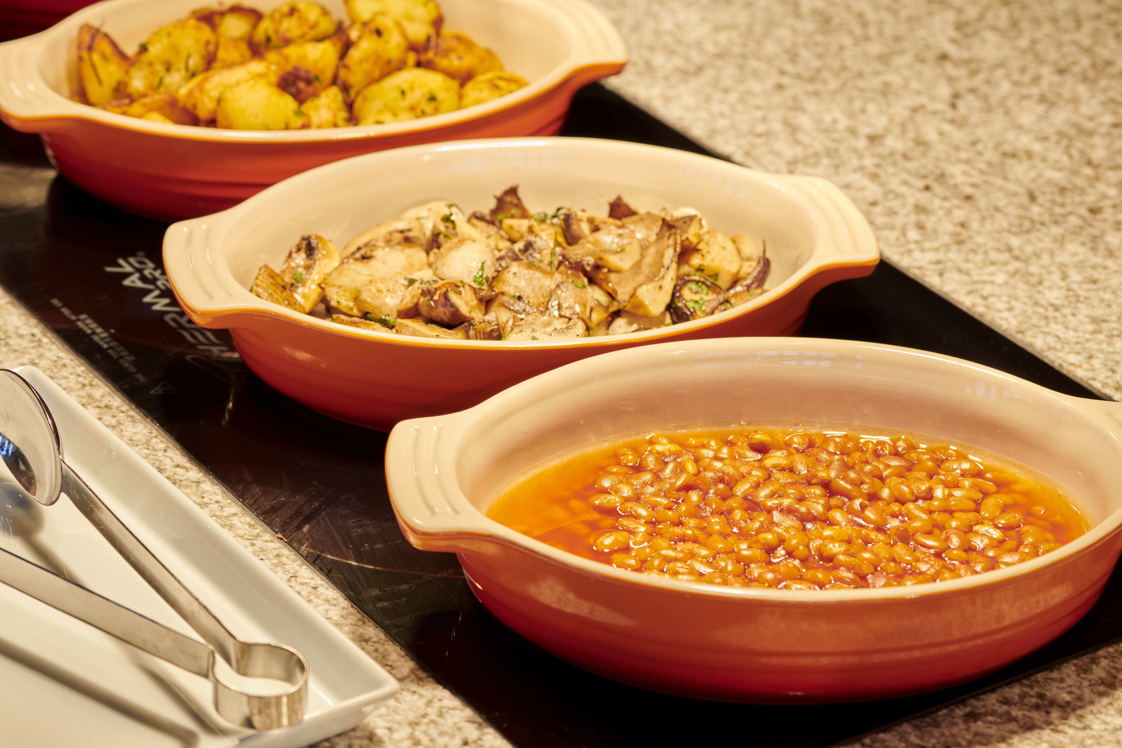 Three ceramic dishes on a stovetop, one with baked potatoes, one with chicken, and one with beans.