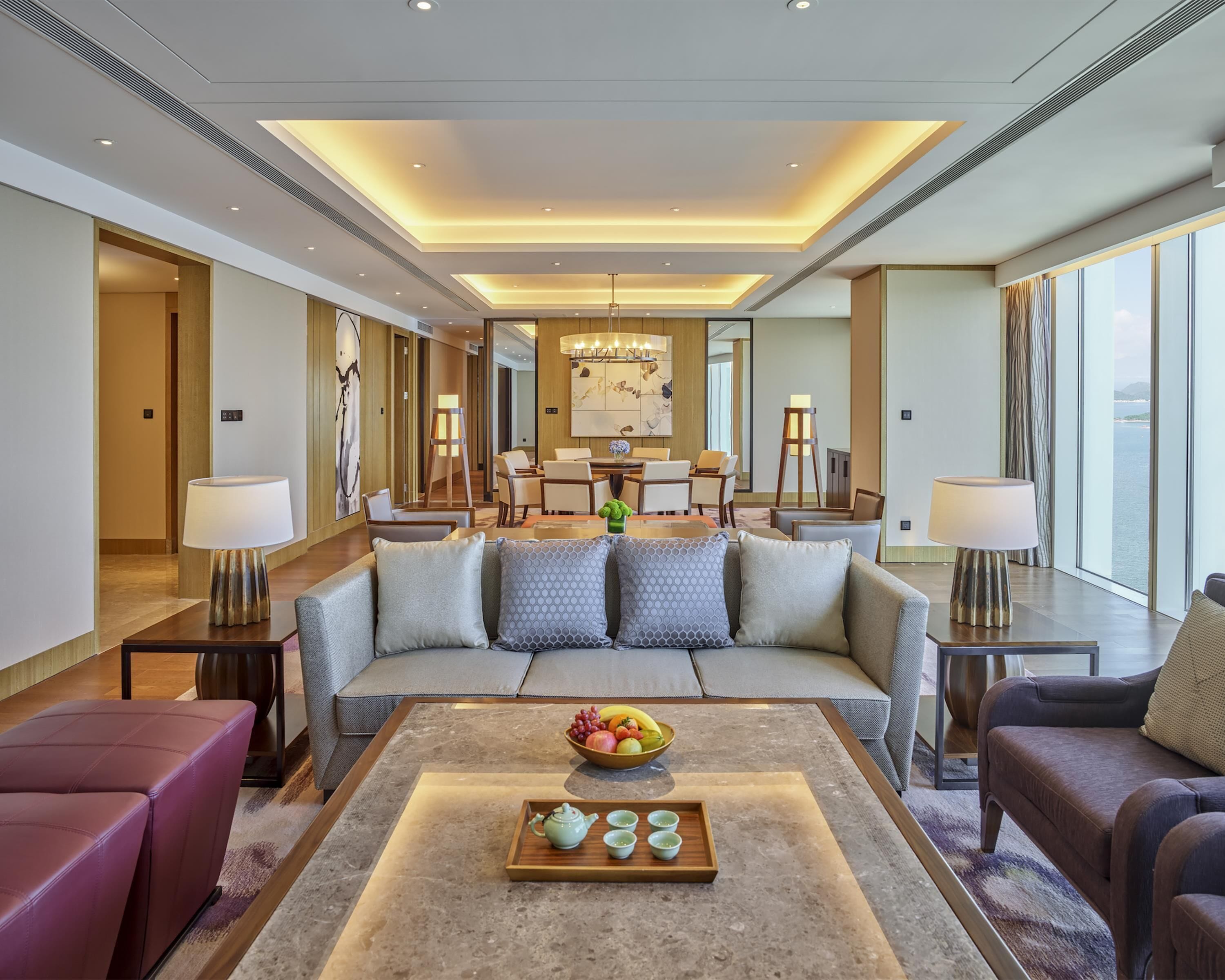 A spacious hotel lobby with a modern design, featuring a central seating area with a sofa and armchairs, a dining table with chairs, and a large window offering a view of the sea.