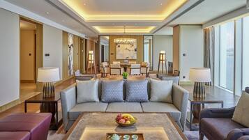 A spacious hotel lobby with a modern design, featuring a central seating area with a sofa and armchairs, a dining table with chairs, and a large window offering a view of the sea.