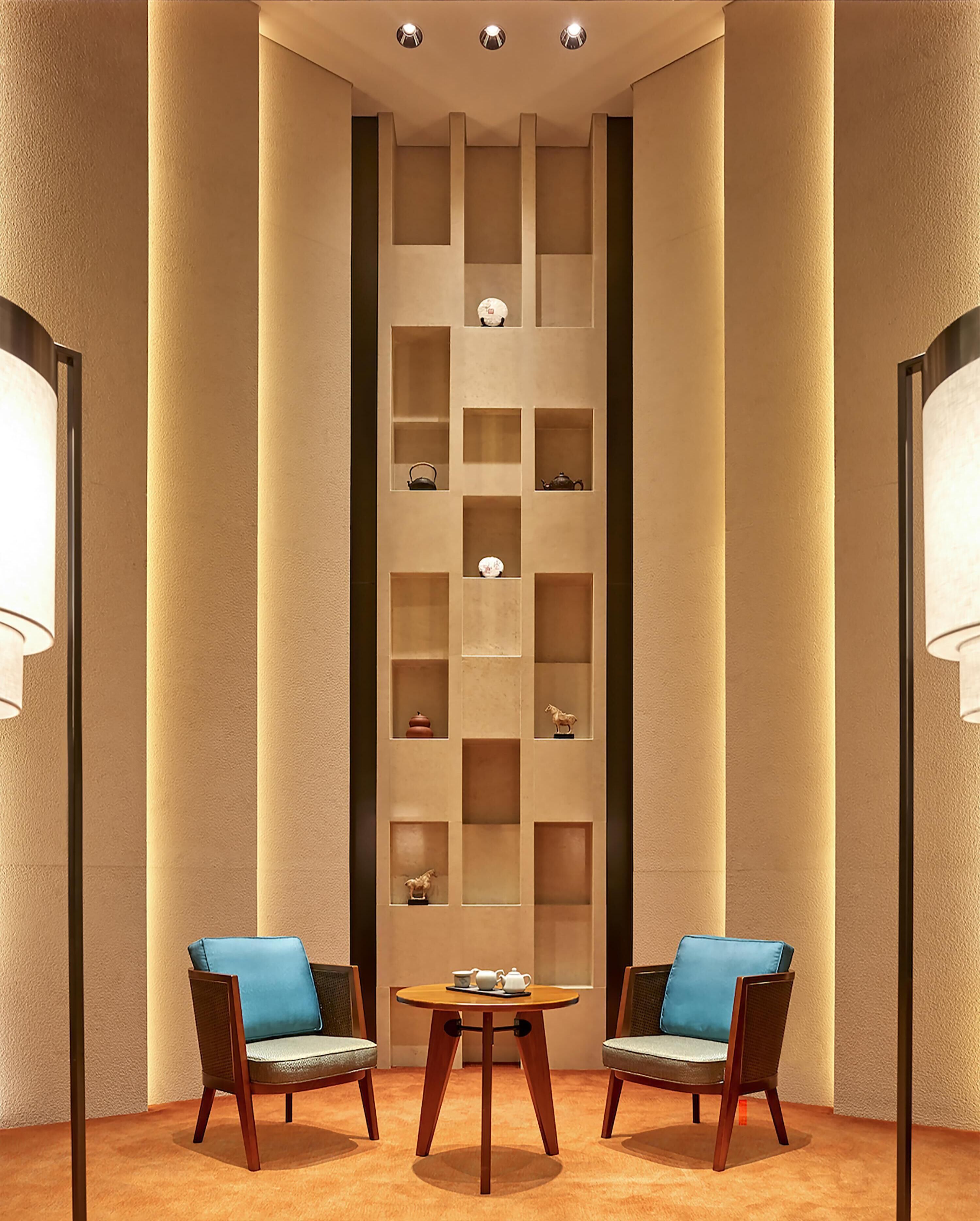 A room with a central table and two chairs, a wall with multiple shelves, and two floor lamps.