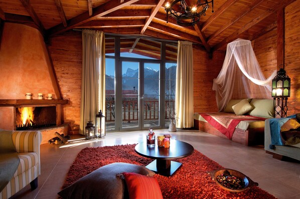A cozy log cabin interior with a fireplace, a comfortable seating area, and a view of mountains through large windows.