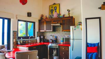 Una cocina con encimera roja, estufa, refrigerador y ventana con vista a palmeras.