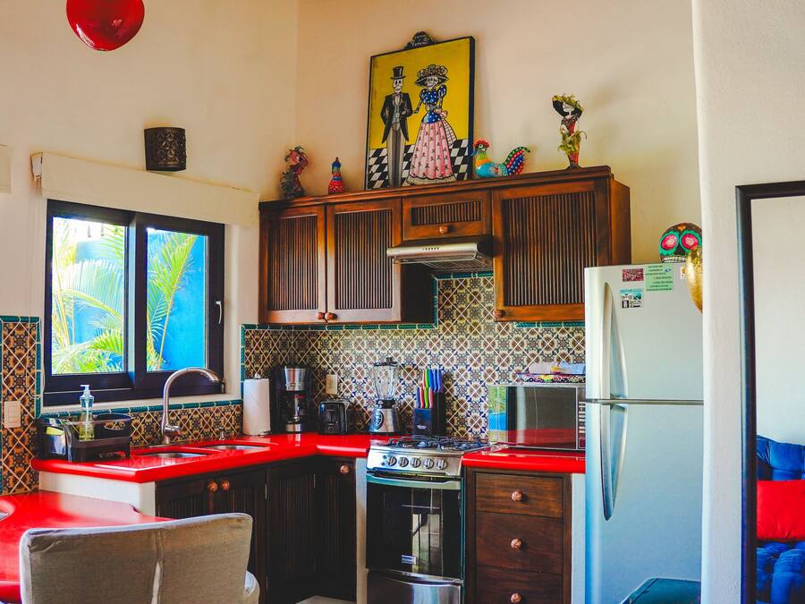 Una cocina con encimera roja, estufa, refrigerador y ventana con vista a palmeras.