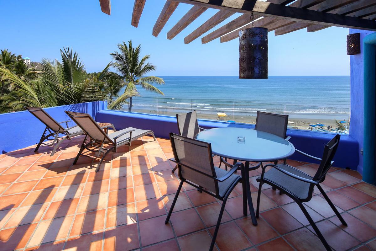 Une terrasse avec une table et des chaises, donnant sur une plage et l’océan.