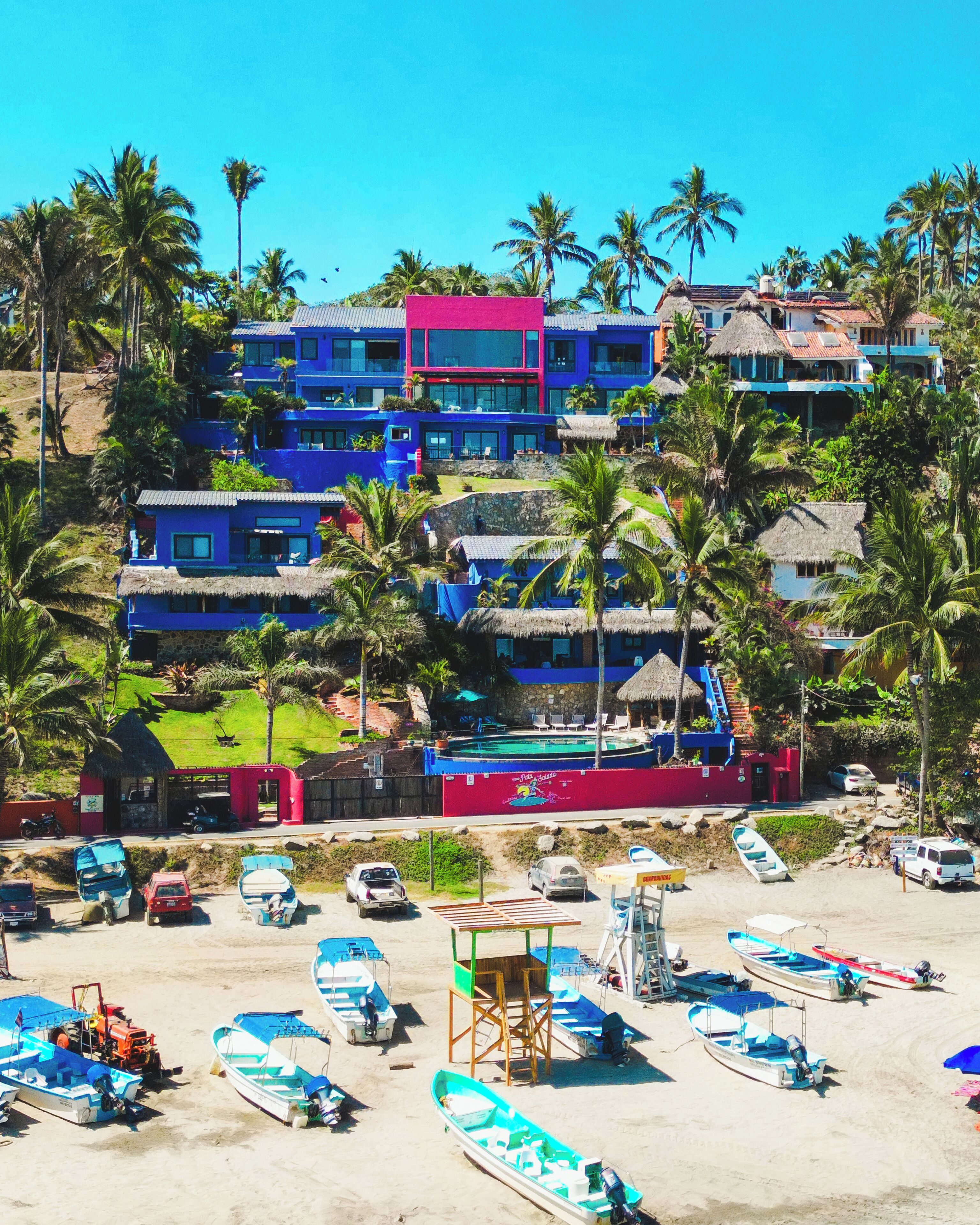 A beachfront property with multiple colorful buildings, a sandy beach with several boats, and palm trees.
