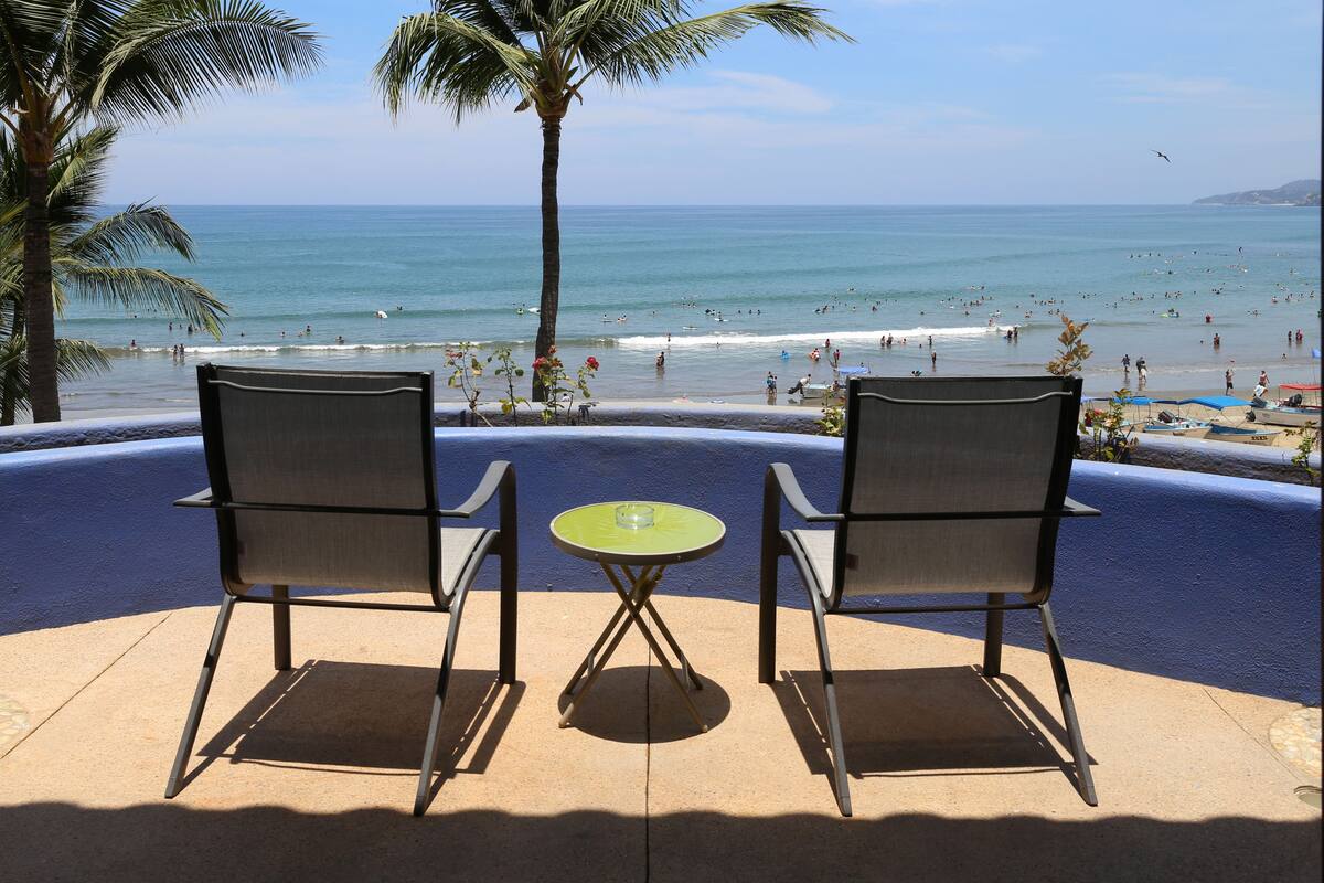 Une terrasse avec deux chaises et une table, donnant sur une plage bordée de palmiers et où l’on peut voir des gens dans l’eau.