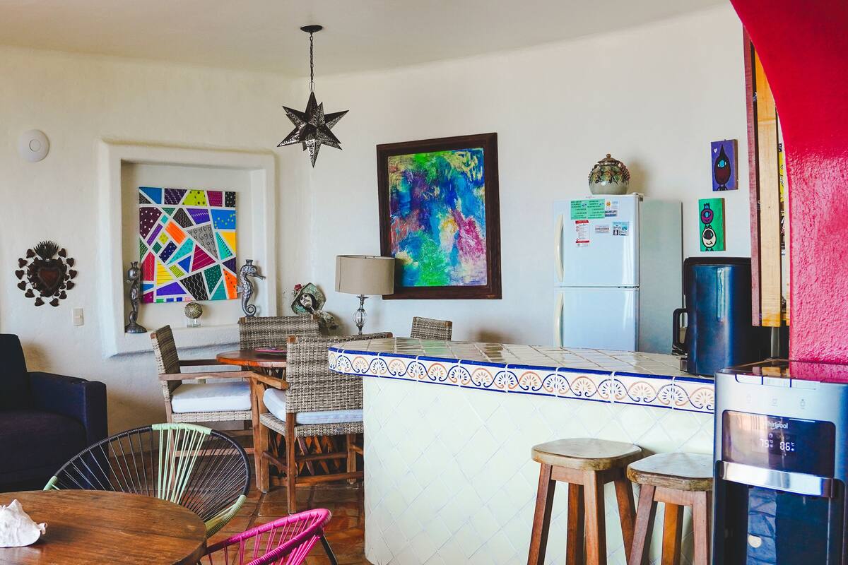 Un salon chaleureux avec un canapé, un coin repas avec des chaises, une cuisine équipée d’un réfrigérateur et une peinture abstraite aux couleurs vives accrochée au mur.