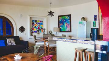 A cozy living room with a sofa, a dining area with chairs, a kitchen with a refrigerator, and a colorful abstract painting on the wall.