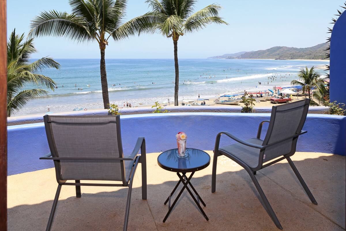 Une terrasse avec deux chaises et une table, donnant sur une plage bordée de palmiers et offrant une vue dégagée sur l’océan.