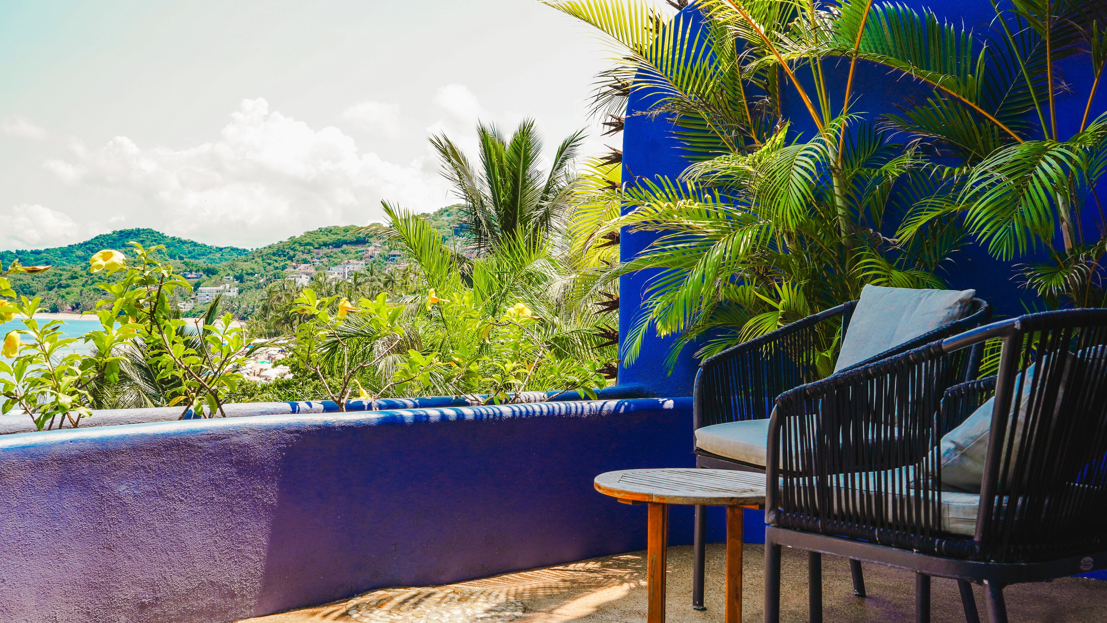 A balcony with a view of a beach and mountains, featuring a blue wall, a wicker chair, and a small table.