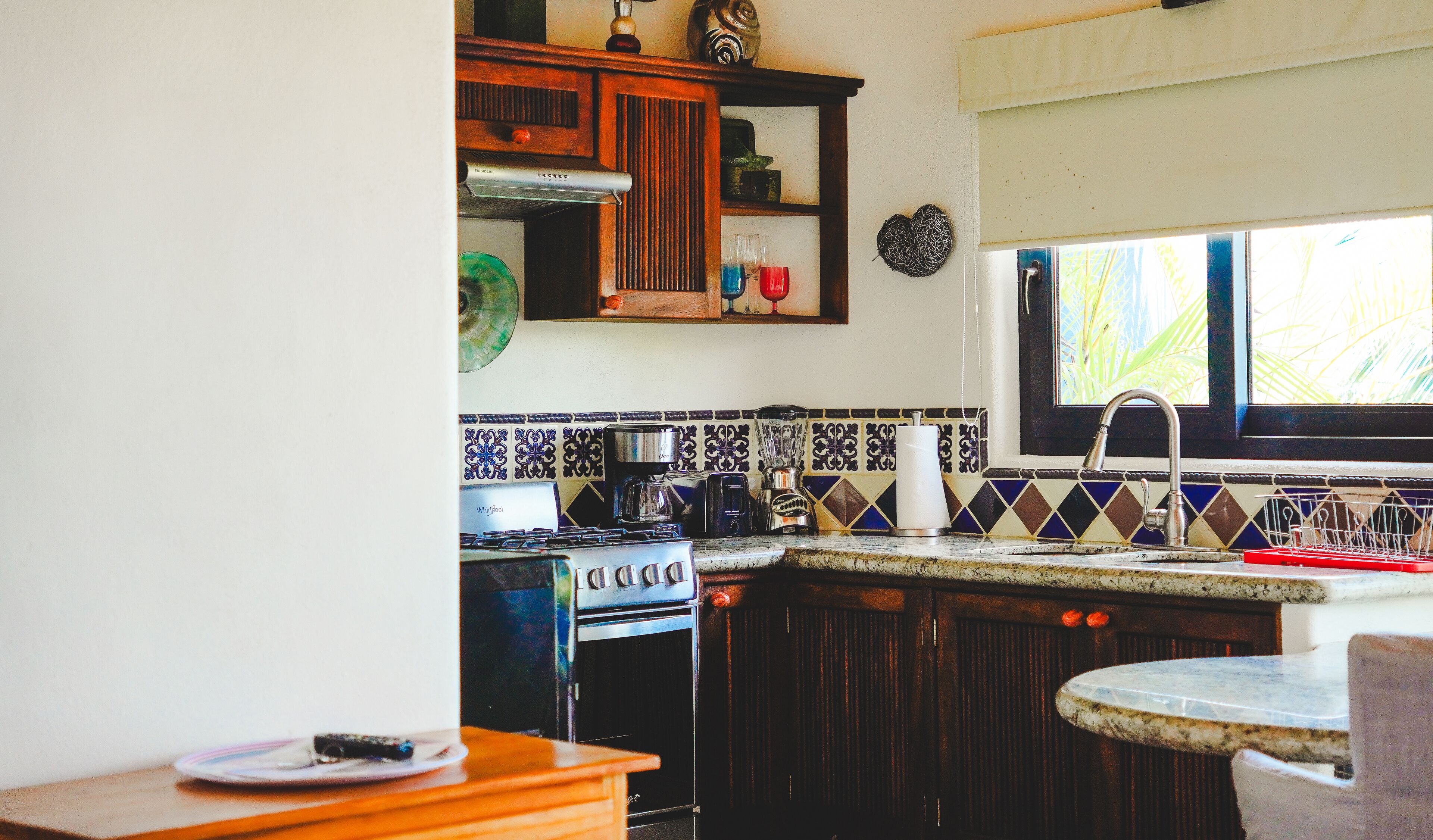 A compact kitchen with a stove, coffee maker, and blender. There is a window with a view of greenery outside.