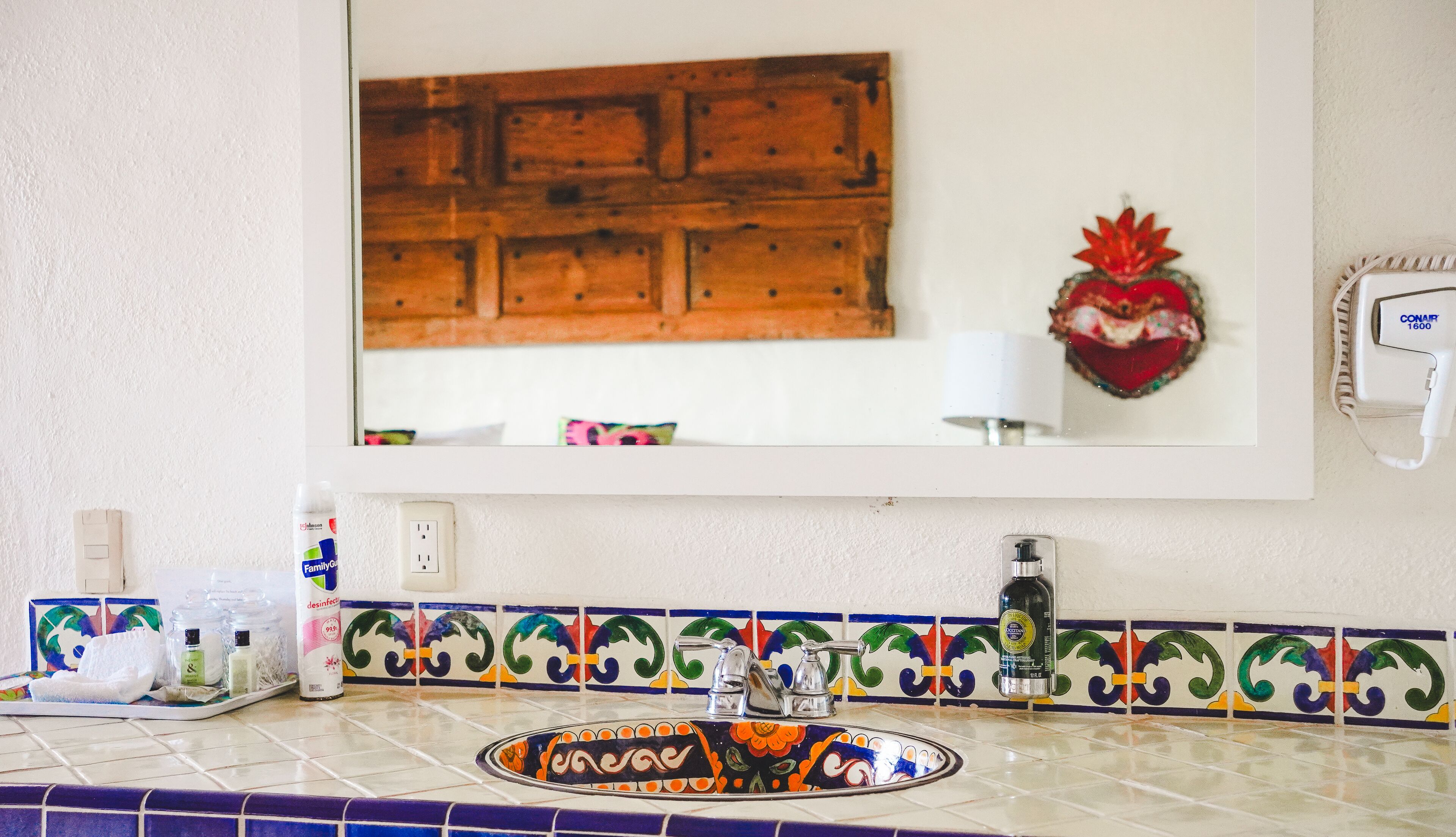A bathroom with a mirror, a sink with a decorative tile backsplash, a soap dispenser, and a bottle of hand sanitizer.